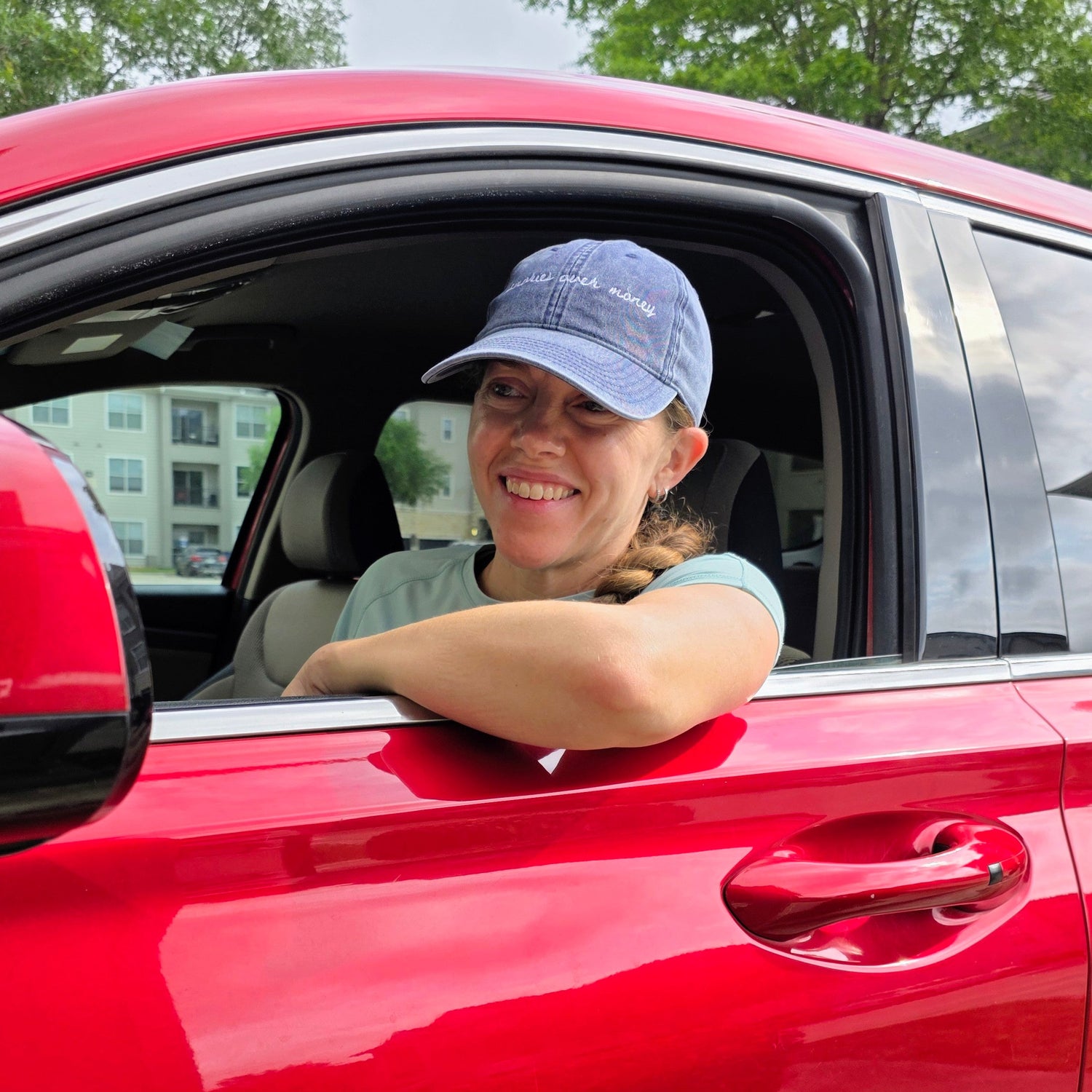 Person sitting in a red car with blue travel hat with "memories over money"  on