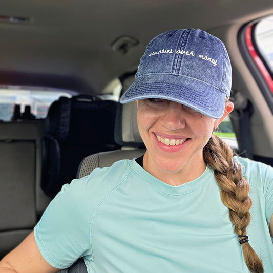 Person wearing a blue travel cap with text "memories over money" inside a car