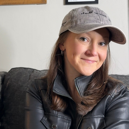 Woman wearing a travel themed hat that says "should we move here" and leather jacket sitting on a couch with framed pictures in the background