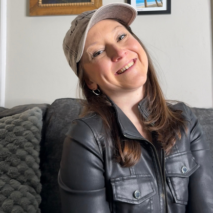 Woman wearing a cap that says "should we move here" and leather jacket sitting on a couch with framed pictures on the wall.