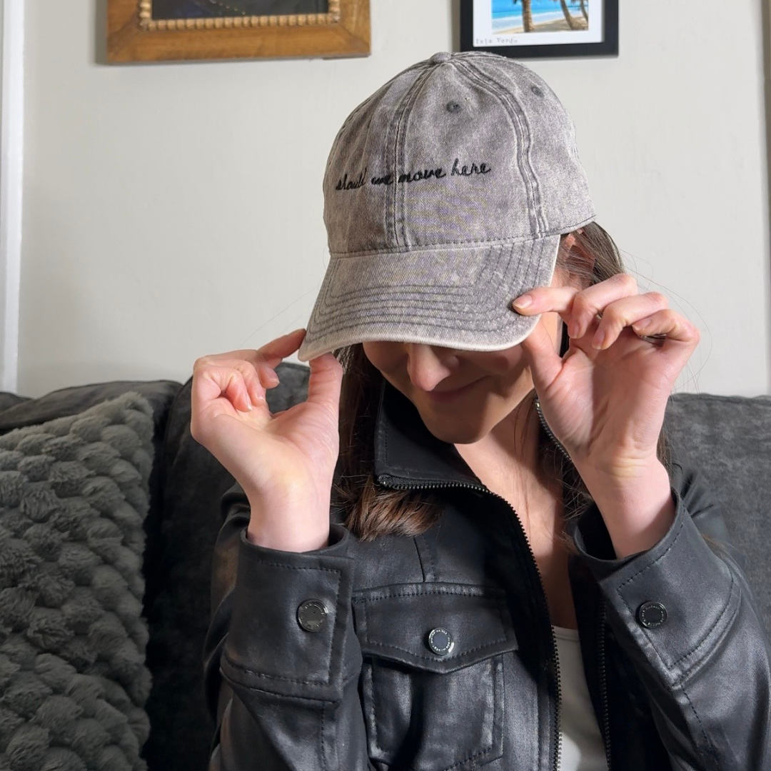 Person wearing a gray baseball cap with text "should we move here", sitting on a couch with framed pictures on the wall.