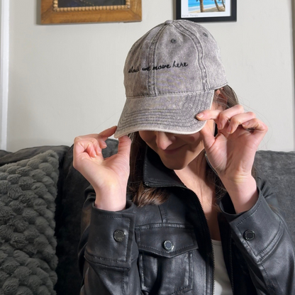 Person wearing a gray baseball cap with text "should we move here", sitting on a couch with framed pictures on the wall.