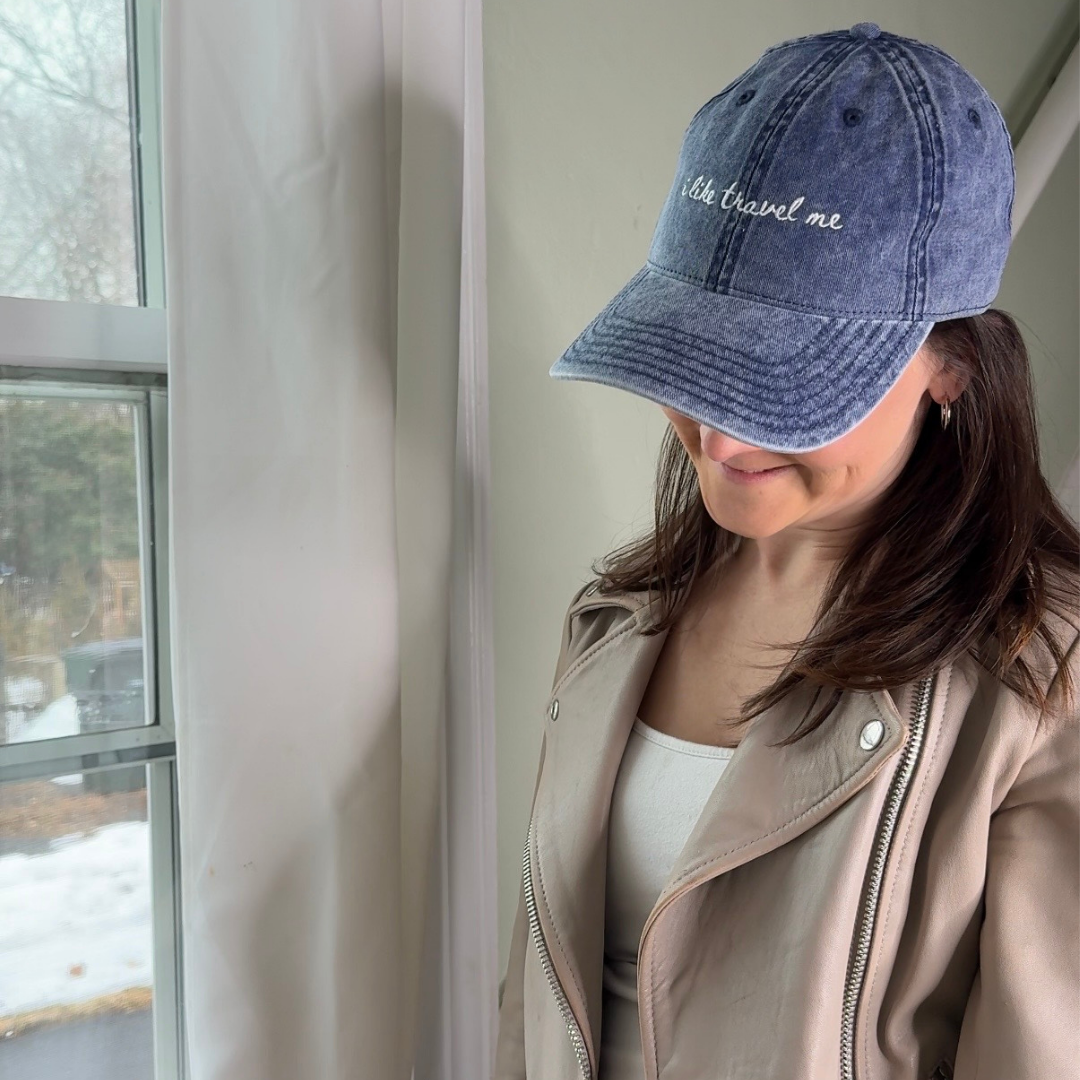 Person wearing a blue denim travel themed hat with "I like travel me" and beige jacket indoors near a window.