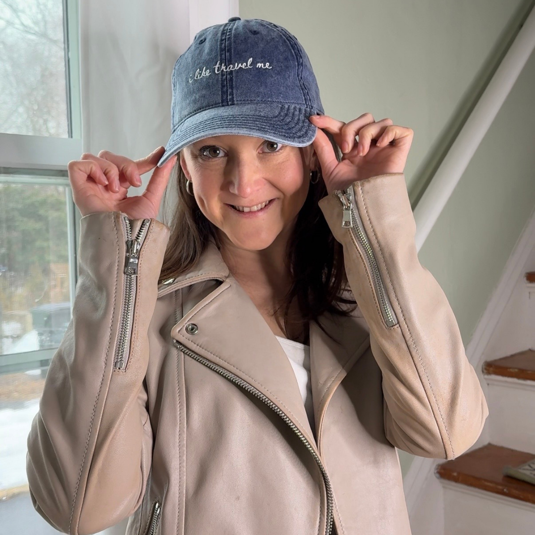 Woman wearing a blue cap travel themed hat with "I like travel me" and beige jacket indoors