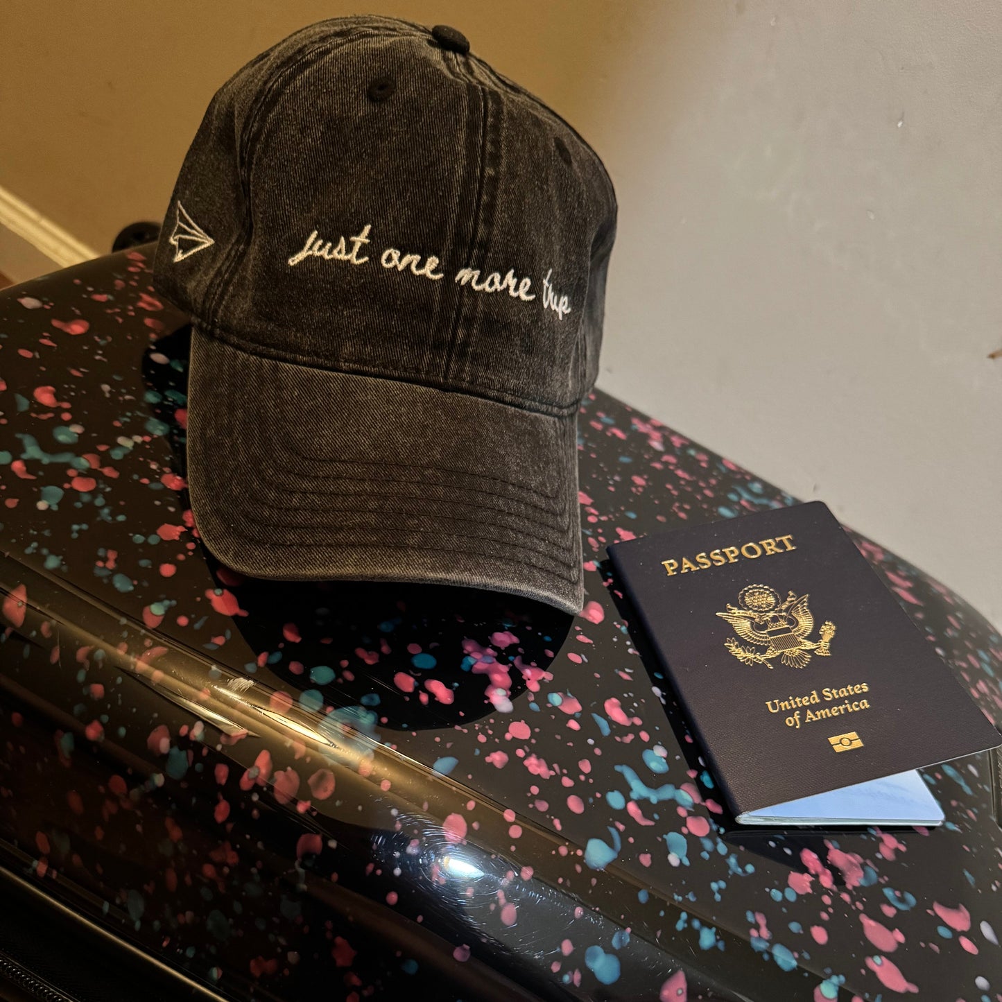 Black travel cap with text "just one more trip", passport, and black suitcase with colorful speckles on a neutral background