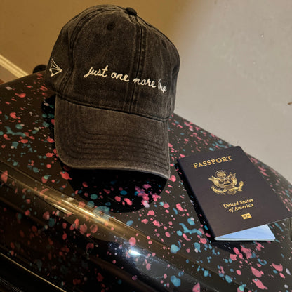 Black travel cap with text "just one more trip", passport, and black suitcase with colorful speckles on a neutral background
