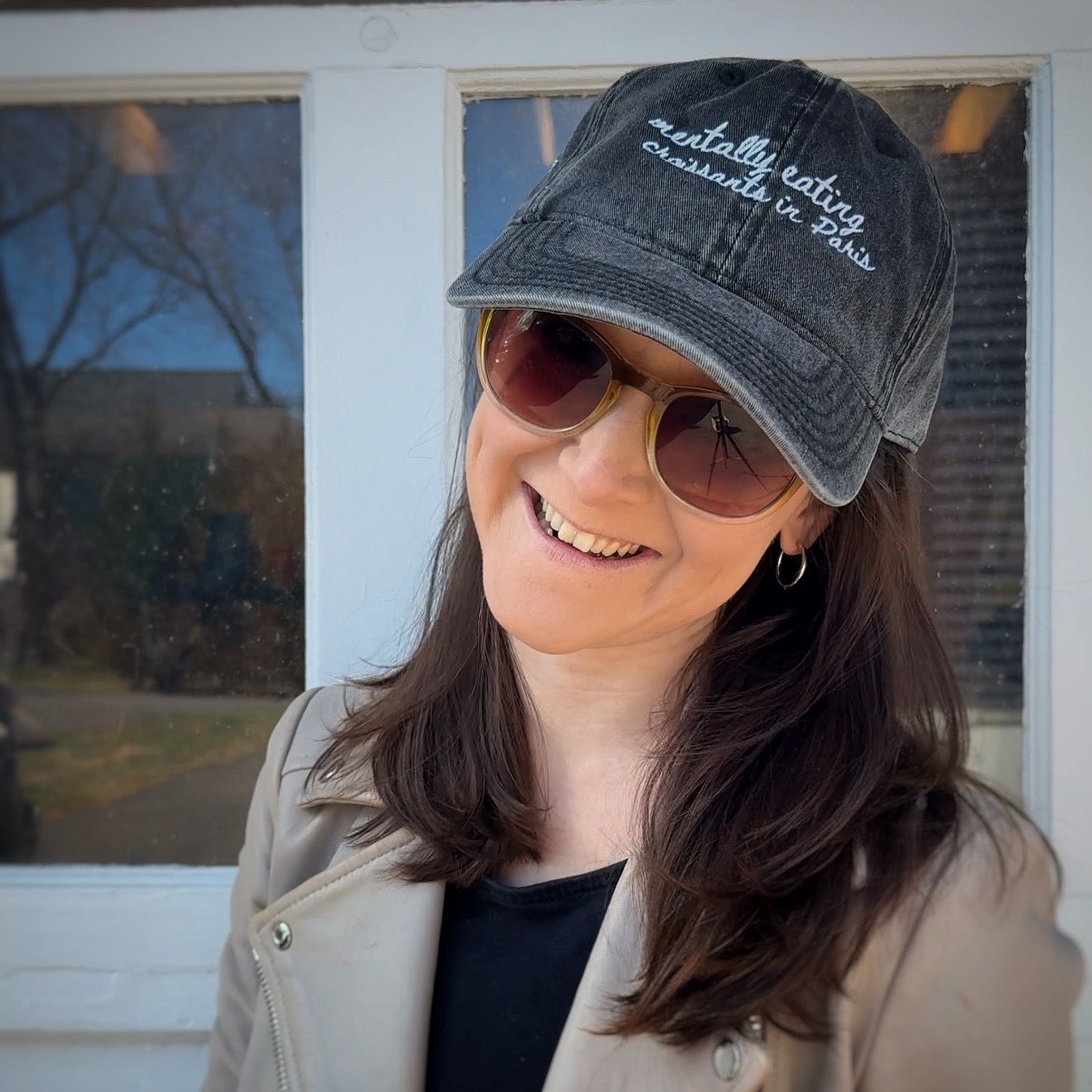 Woman wearing a travel hat with "mentally eating croissants in Paris" and sunglasses, standing in front of a door.