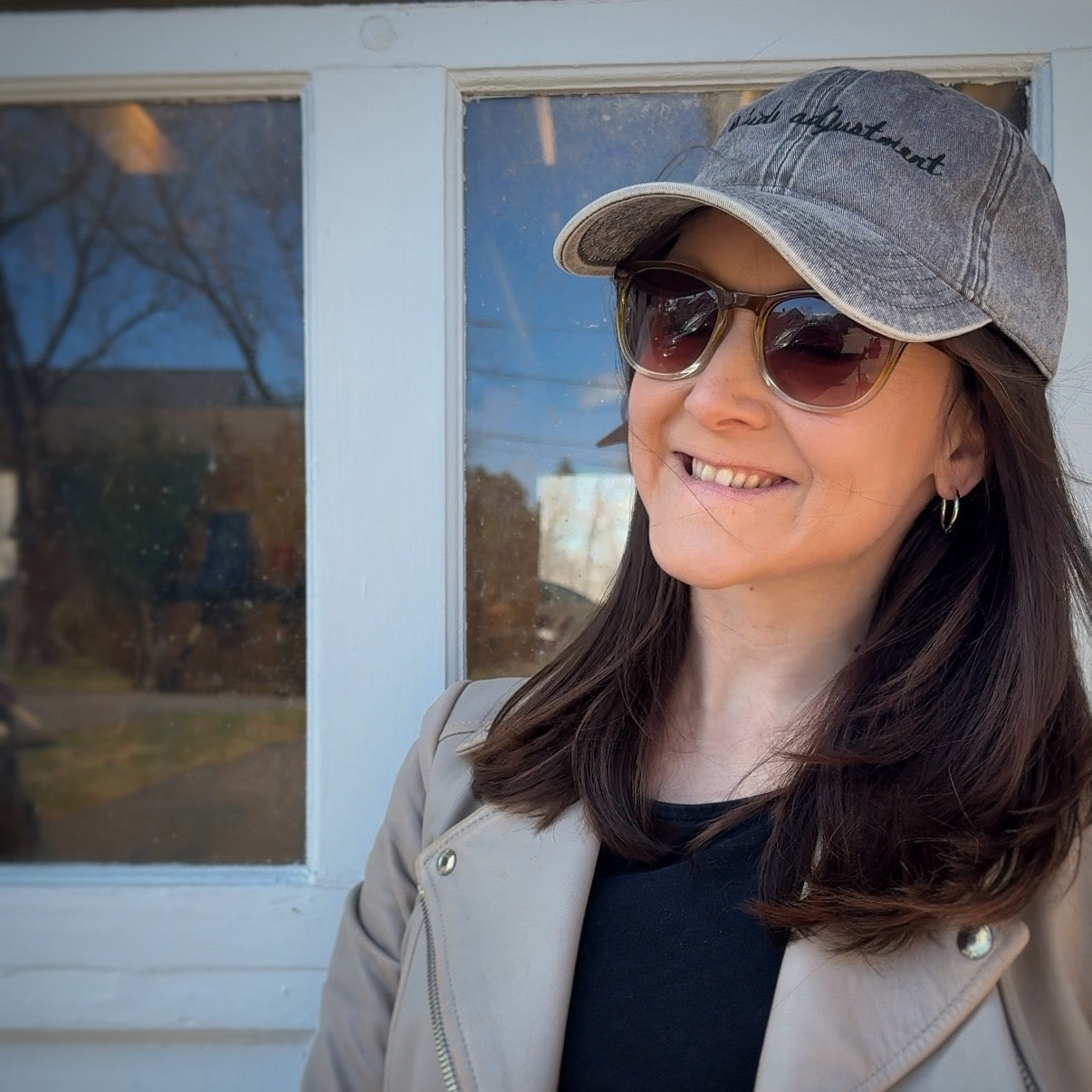 Woman wearing a travel themed hat with words "altitude adjustment" and sunglasses standing in front of a window