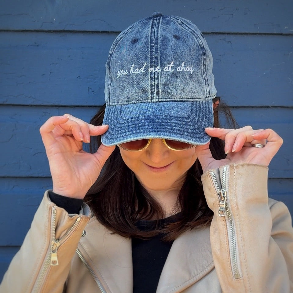 Person wearing a denim travel themed hat with "you had me at ahoy", sunglasses, and a beige jacket against a blue wall.