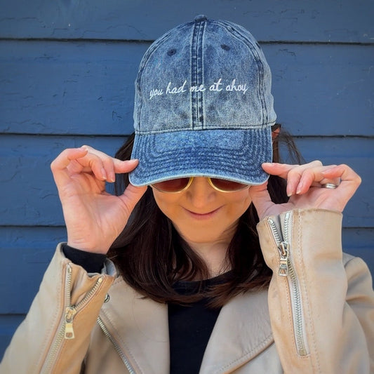 Person wearing a denim travel themed hat with "you had me at ahoy", sunglasses, and a beige jacket against a blue wall.