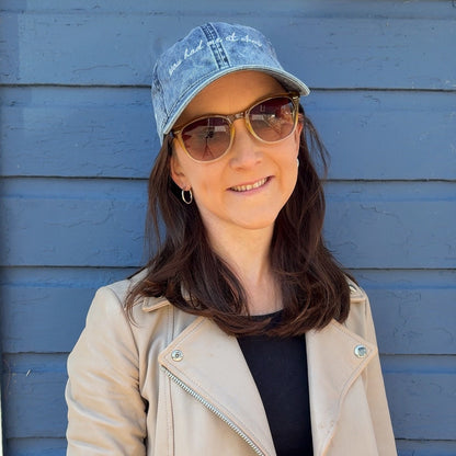 Woman wearing a beige jacket, sunglasses, and a blue cap that says "you had me at ahoy" against a blue wall.