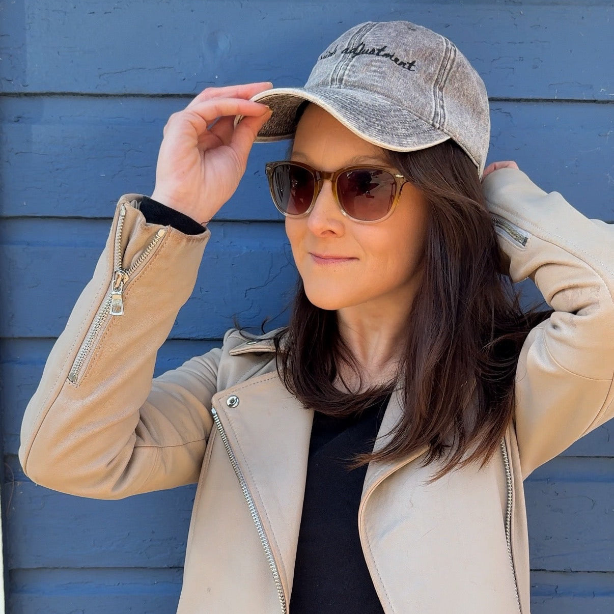 Woman wearing a grey travel themed hat with the words "altitude adjustment"  , sunglasses, and jacket against a blue wall