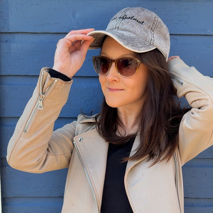 Woman wearing a grey travel themed hat with the words "altitude adjustment"  , sunglasses, and jacket against a blue wall
