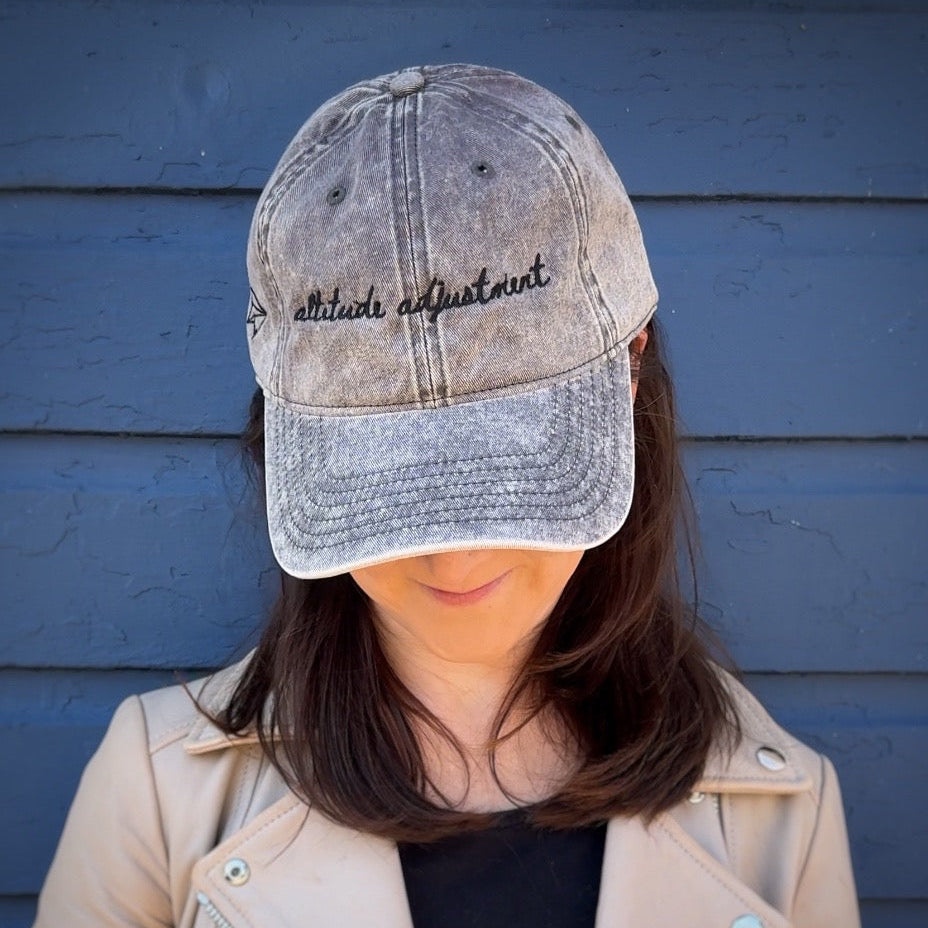 Person wearing a distressed baseball cap with text "altitude adjustment"  against a blue wall