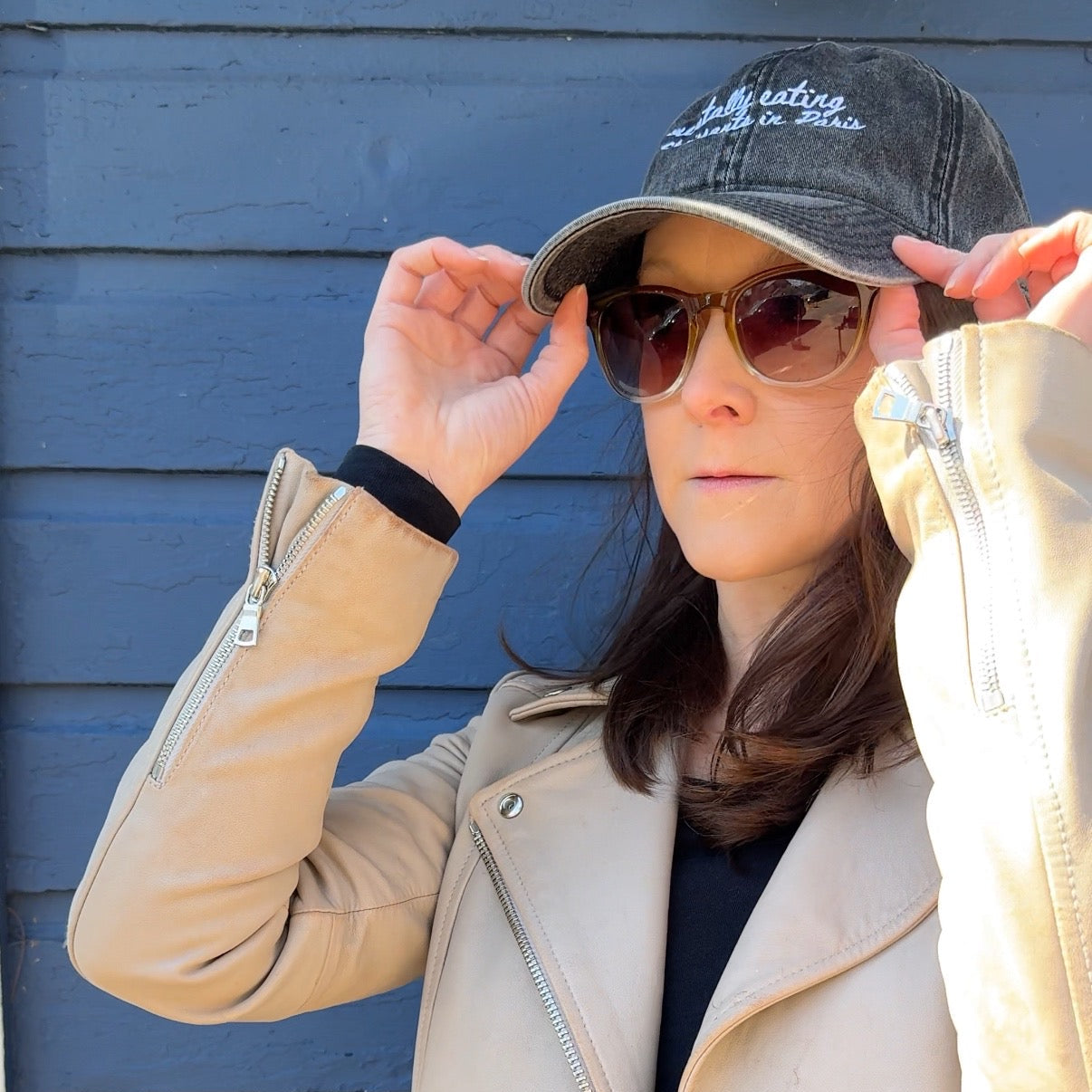 Person wearing a beige jacket, sunglasses, and a travel hat with "mentally eating croissants in Paris" against a blue wall.
