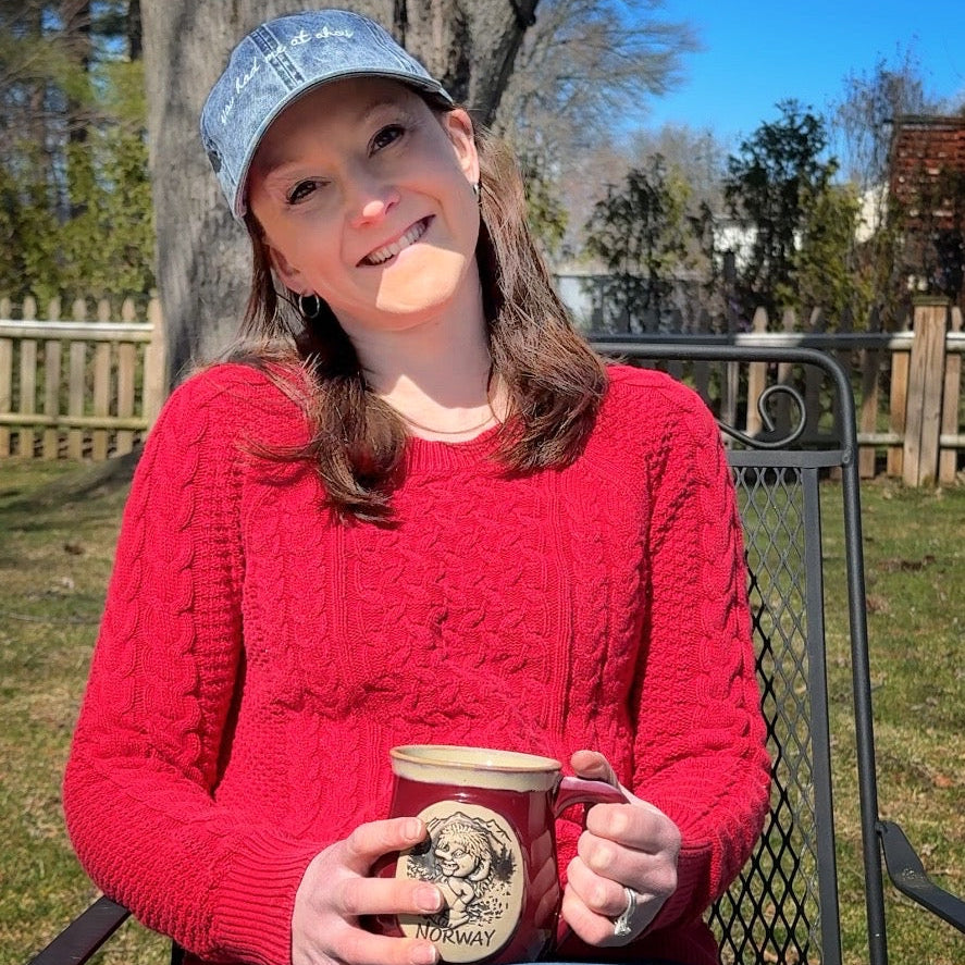 Person wearing a red sweater and blue cap that says "you had me at ahoy", holding a cup outdoors.