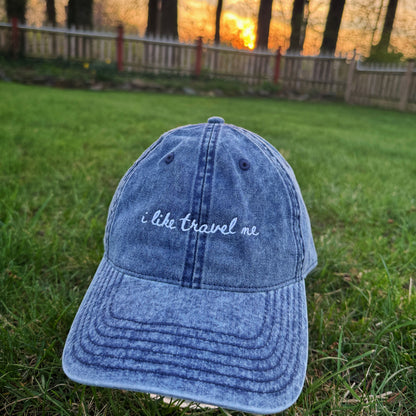 Blue denim cap with 'i like travel me' text on grass at sunset
